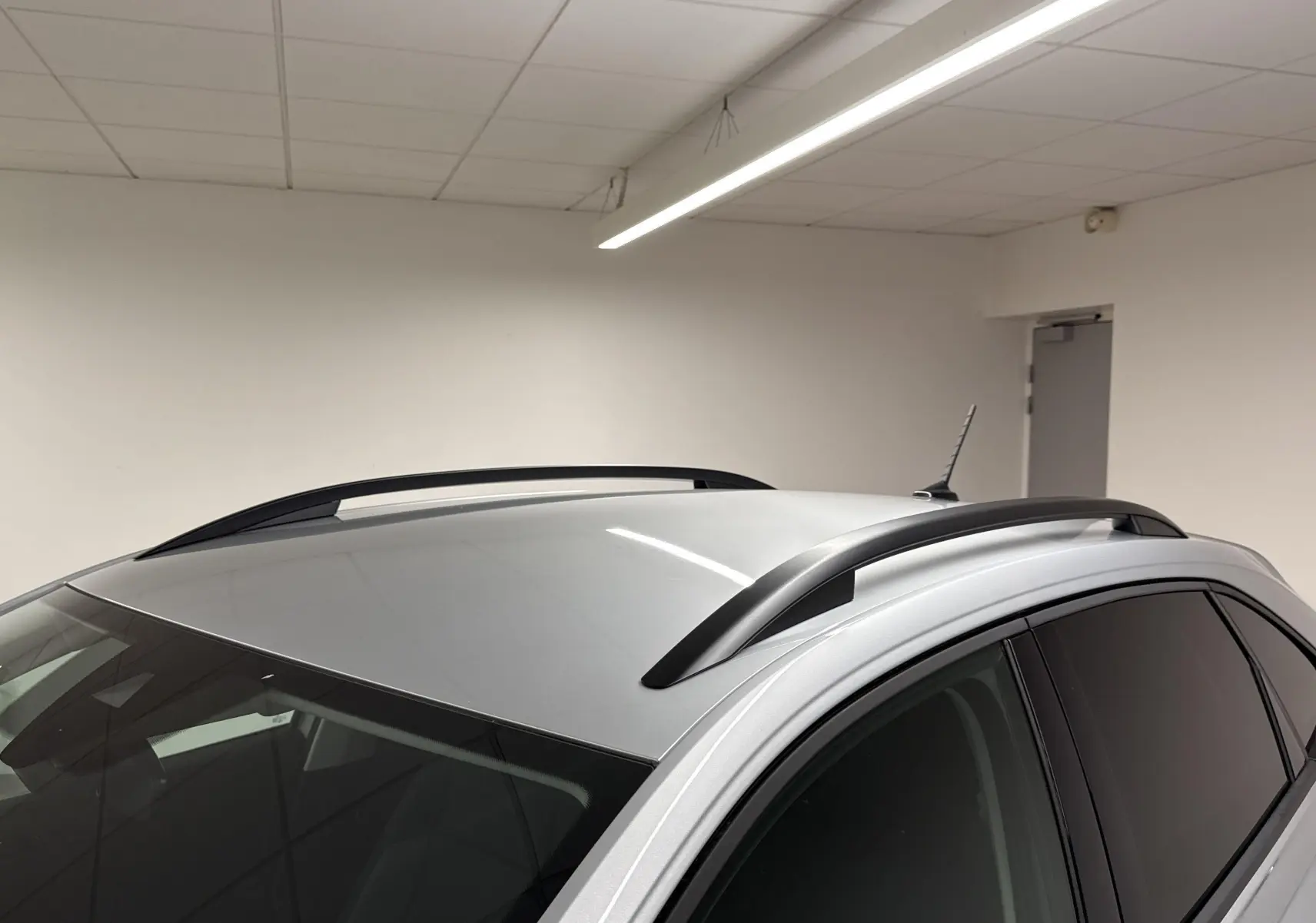 Vue 3/4 arrière du Volkswagen Taigo gris cendré 2024 avec barres de toit noires dans un garage blanc.