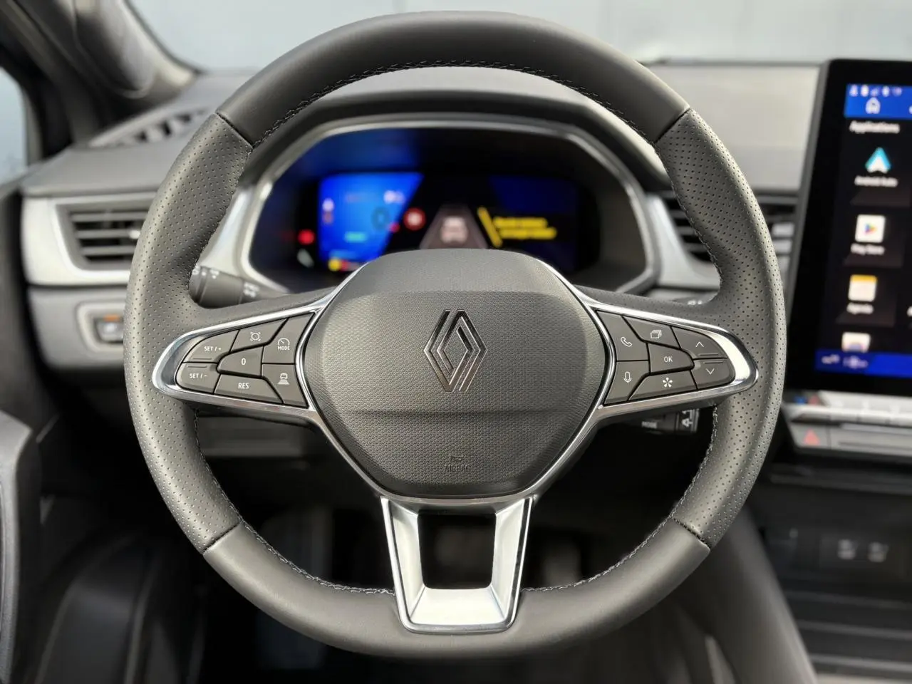 Volant en cuir noir avec commandes intégrées et logo Renault au centre, tableau de bord numérique en arrière-plan.