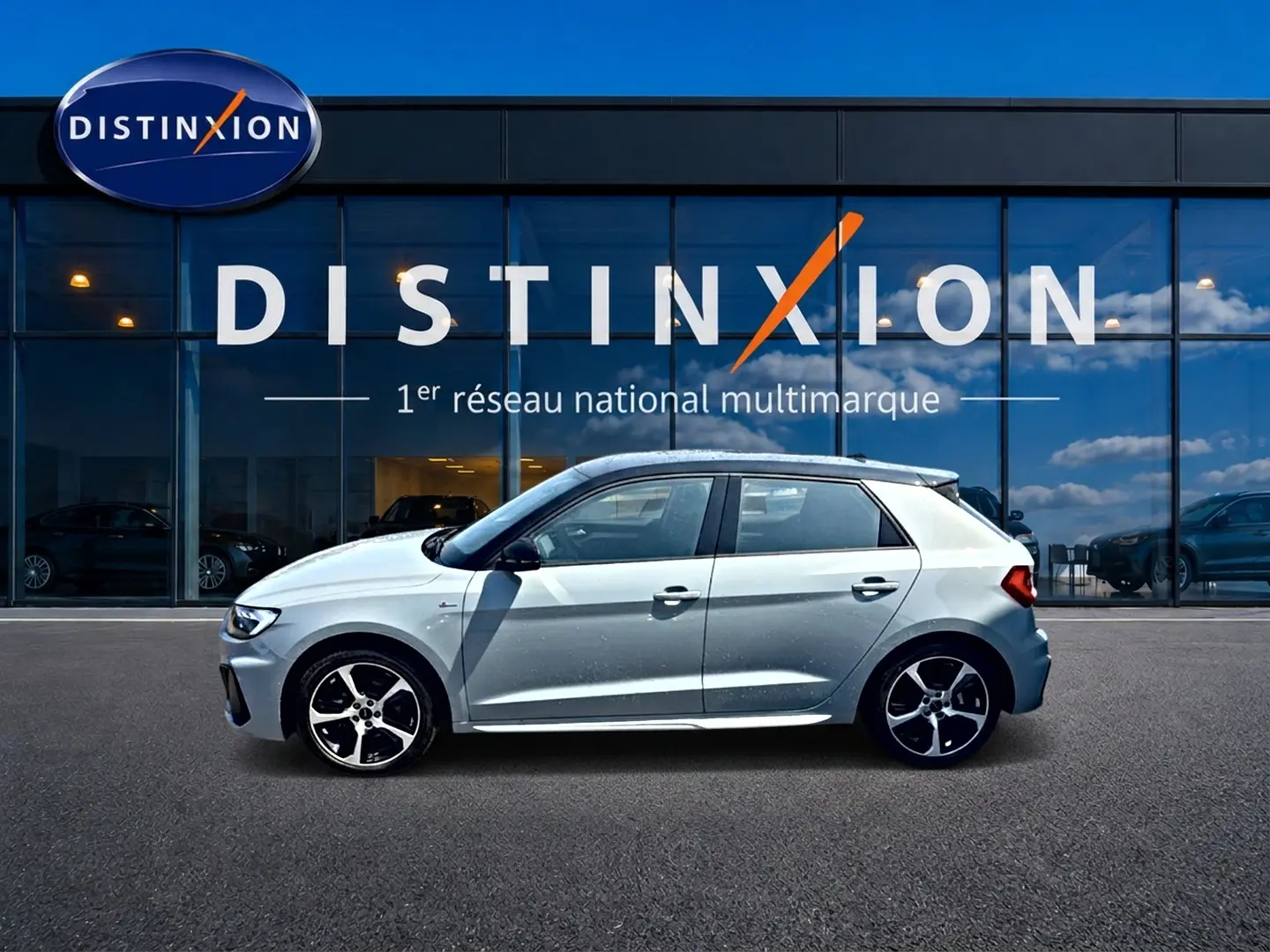 Profil droit d'une Audi A1 Sportback 30 TFSI S line gris arrow avec jantes alliage noires devant une vitrine Distinxion.