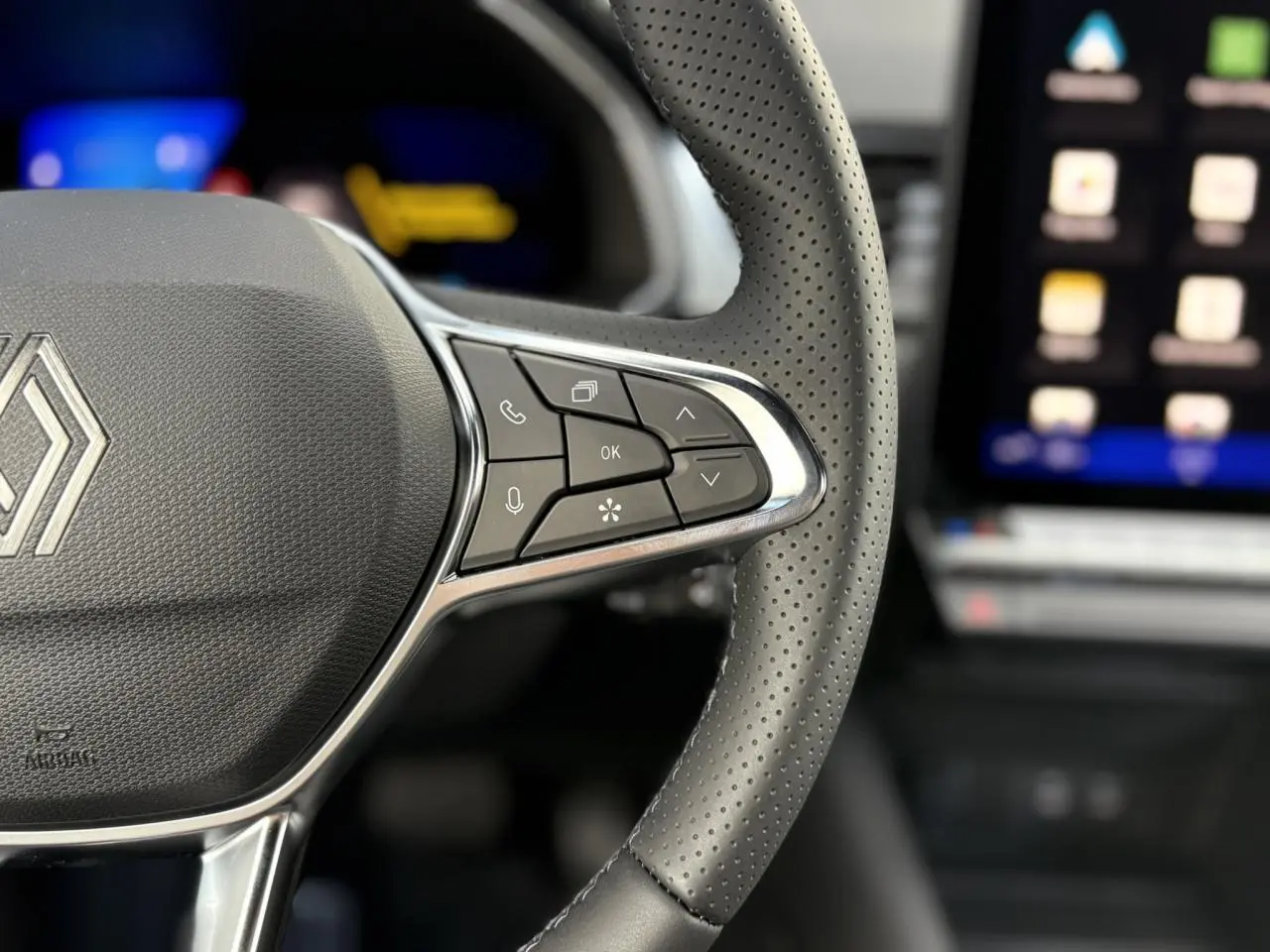 Gros plan sur le volant en cuir perforé du Renault SYMBIOZ 2025 avec commandes multifonctions et écran tactile flou en arrière-plan.