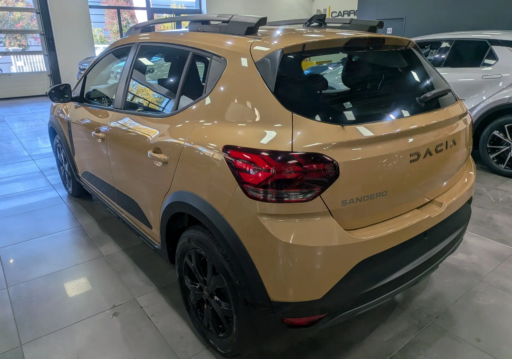 Vue 3/4 arrière droite d'une Dacia Sandero Stepway beige Safari avec jantes noires et barres de toit en showroom.