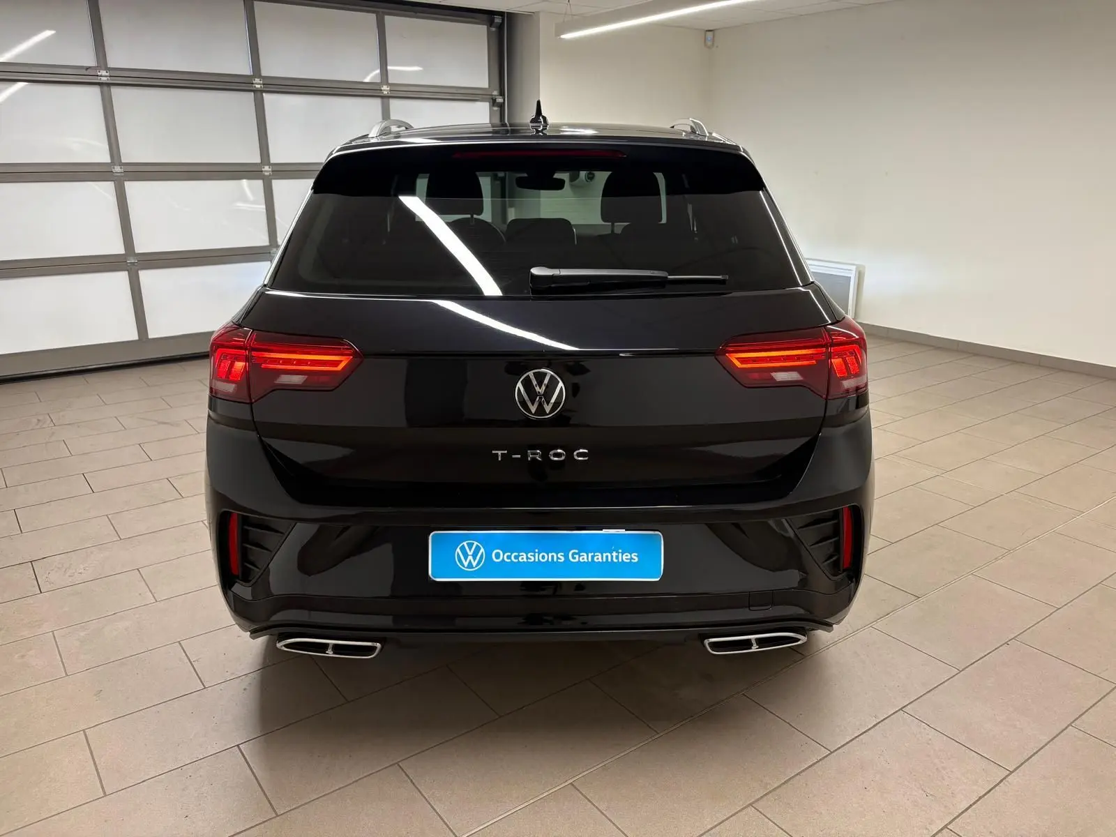 Vue arrière du Volkswagen T-Roc noir intense 2025 avec feux allumés et double sortie d'échappement chromée.