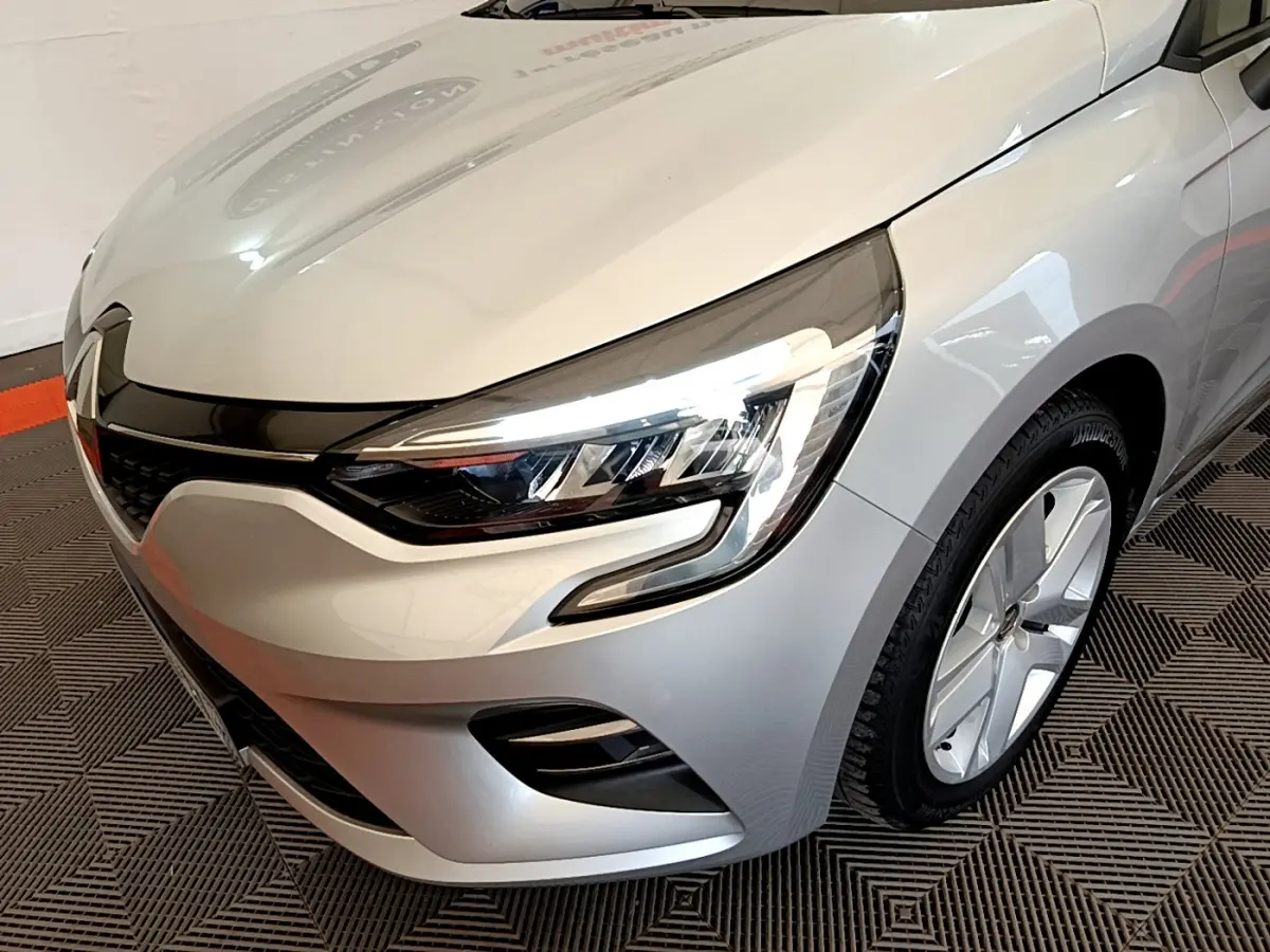 Vue 3/4 avant droit d'une Renault Clio Business TCe 100 gris clair, mettant en valeur ses phares LED et la calandre noire brillante.