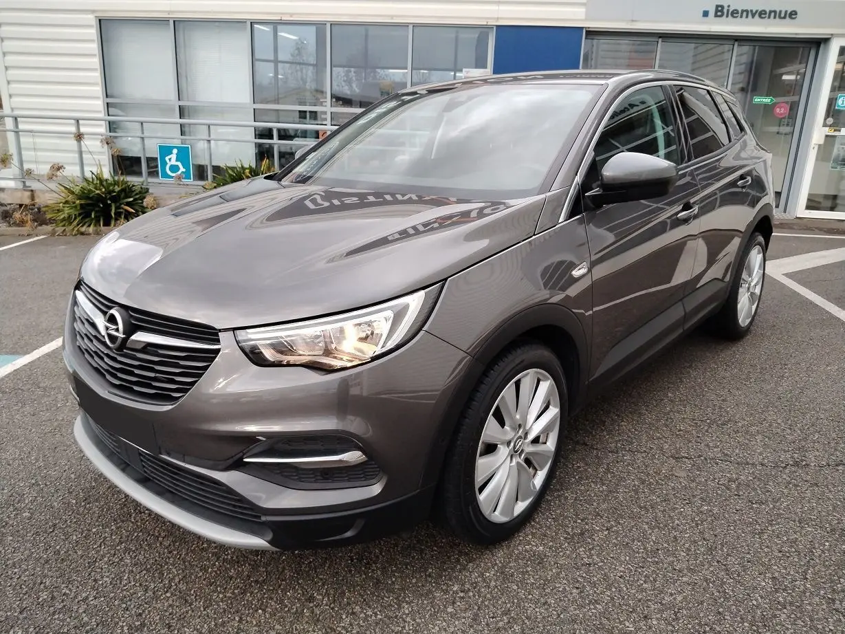 Opel Grandland X gris platinium vu de 3/4 avant droit avec phares allumés sur parking devant bâtiment.