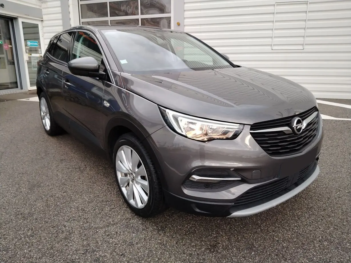 Opel Grandland X gris platinium en 3/4 avant droit, avec phares allumés et jantes alliage visibles.