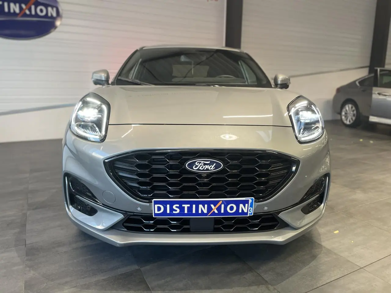 Vue frontale d'un Ford Puma gris clair avec calandre noire hexagonale et phares LED allumés en intérieur showroom.