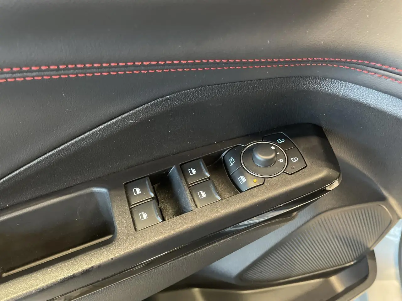 Gros plan sur la commande des vitres et rétroviseurs côté conducteur, avec surpiqûres rouges sur la garniture noire.