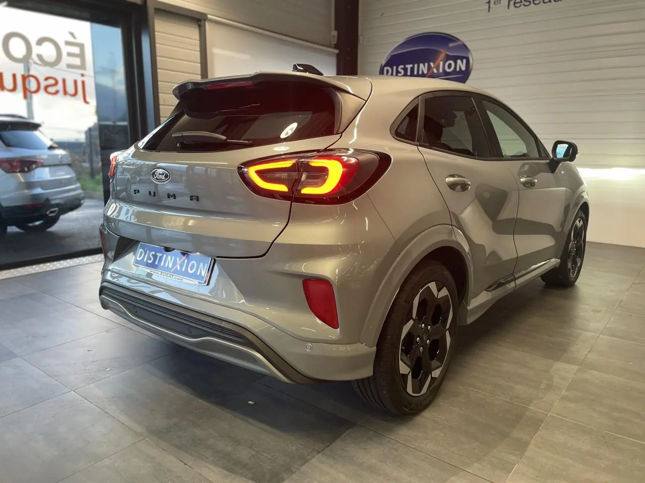 Vue 3/4 arrière droite d'un Ford Puma gris clair avec feux arrière allumés en intérieur showroom.