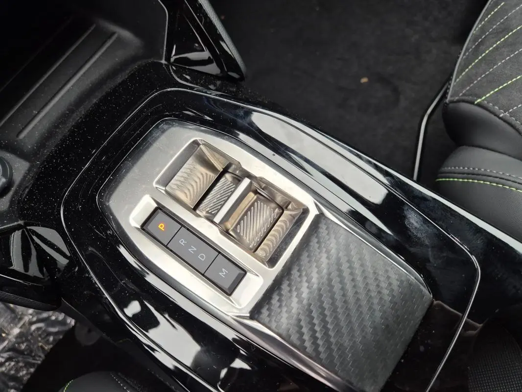 Gros plan sur la console centrale noire brillante de la Peugeot 208 Hybrid 110 e-DCS6 GT avec levier de vitesses électrique et finition carbone.