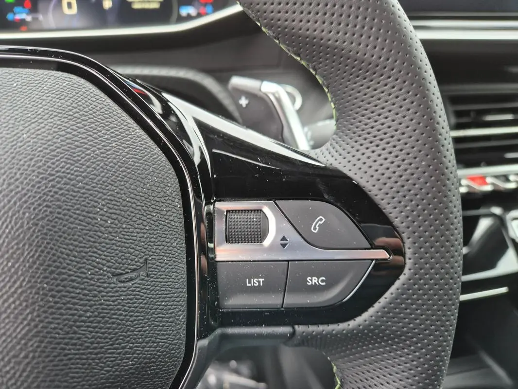 Gros plan sur le volant perforé de la Peugeot 208 Hybrid 110 e-DCS6 GT gris, avec commandes audio et téléphone intégrées.