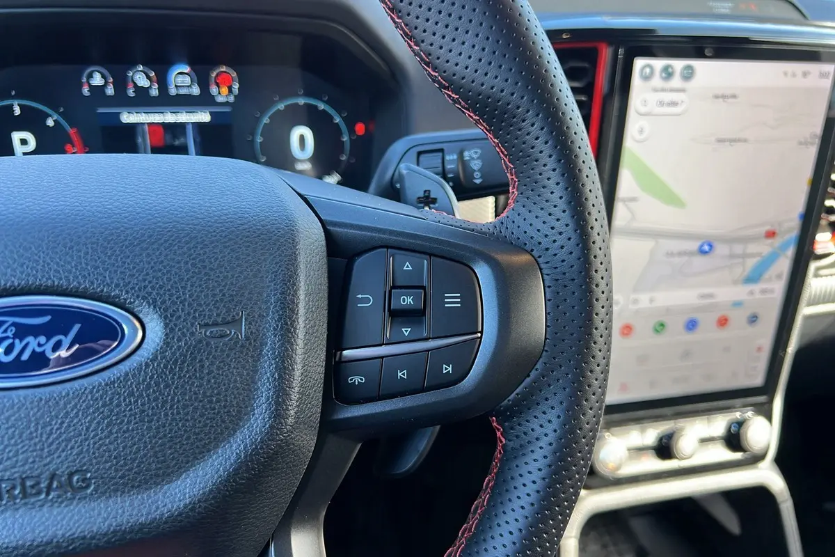 Gros plan sur le volant cuir perforé avec surpiqûres rouges du Ford Ranger Raptor 2025, écran tactile de navigation visible.