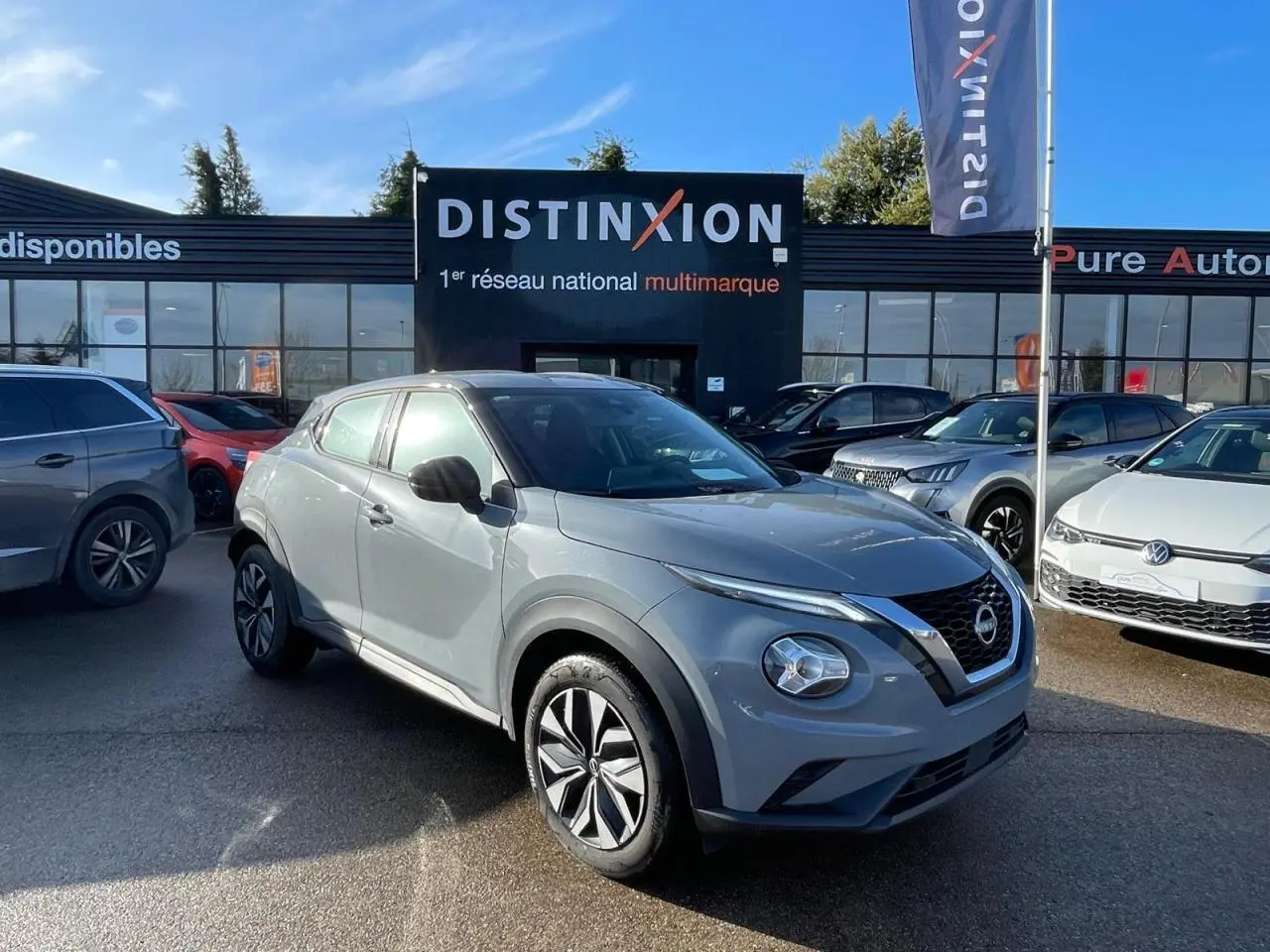 Nissan Juke II gris ceramic en 3/4 avant droit, avec jantes alliage noires et calandre V-motion distinctive.