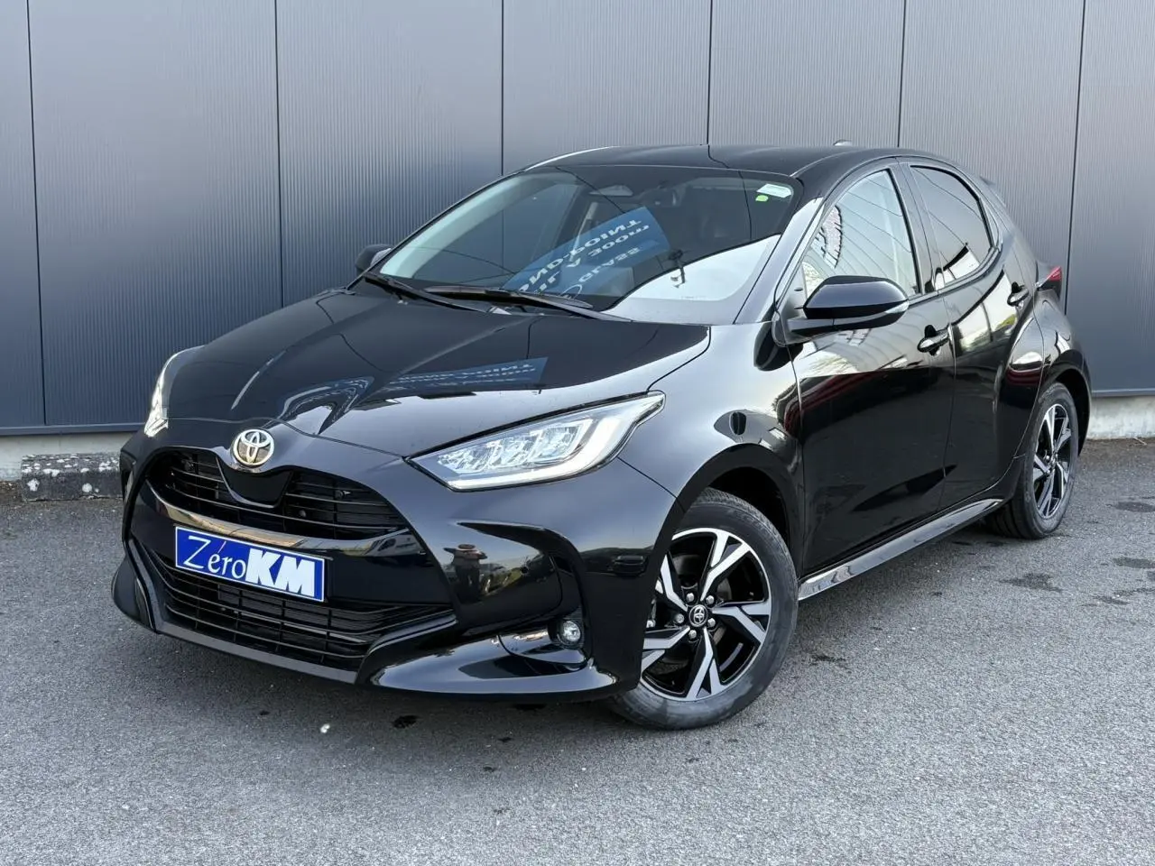Toyota Yaris Hybride 2025 noir intense vue 3/4 avant avec jantes alliage et phares LED allumés.