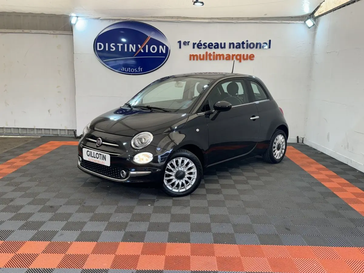 Fiat 500 noire vue en 3/4 avant droit dans un showroom avec jantes alliage et phares ronds allumés.