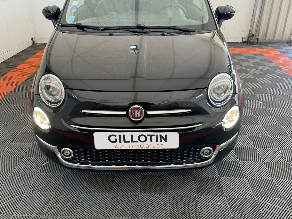 Vue frontale d'une Fiat 500 noire avec phares allumés et calandre chromée dans un garage.