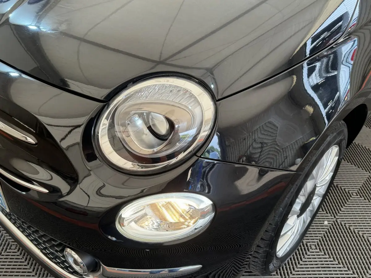 Gros plan sur l'avant droit noir brillant de la Fiat 500 1.2 8V 69ch Lounge avec phare rond et antibrouillard allumé.