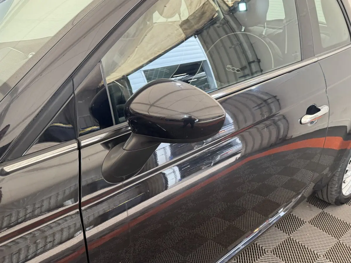 Gros plan sur le rétroviseur côté gauche d'une Fiat 500 noire 2019, avec reflet de l'intérieur et poignée chromée visible.