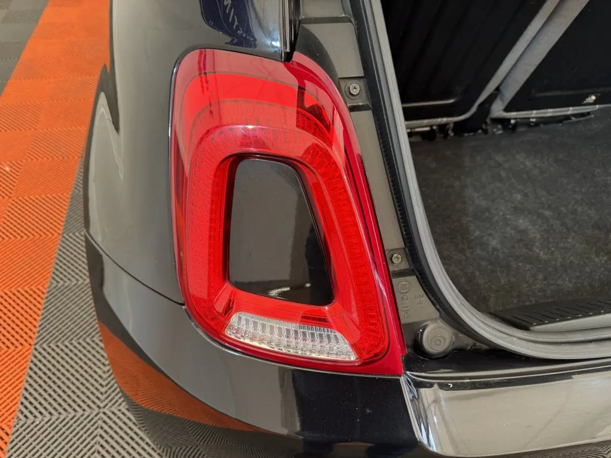 Gros plan sur le feu arrière droit rouge et blanc d'une FIAT 500 noire avec coffre ouvert sur sol carrelé orange et gris.