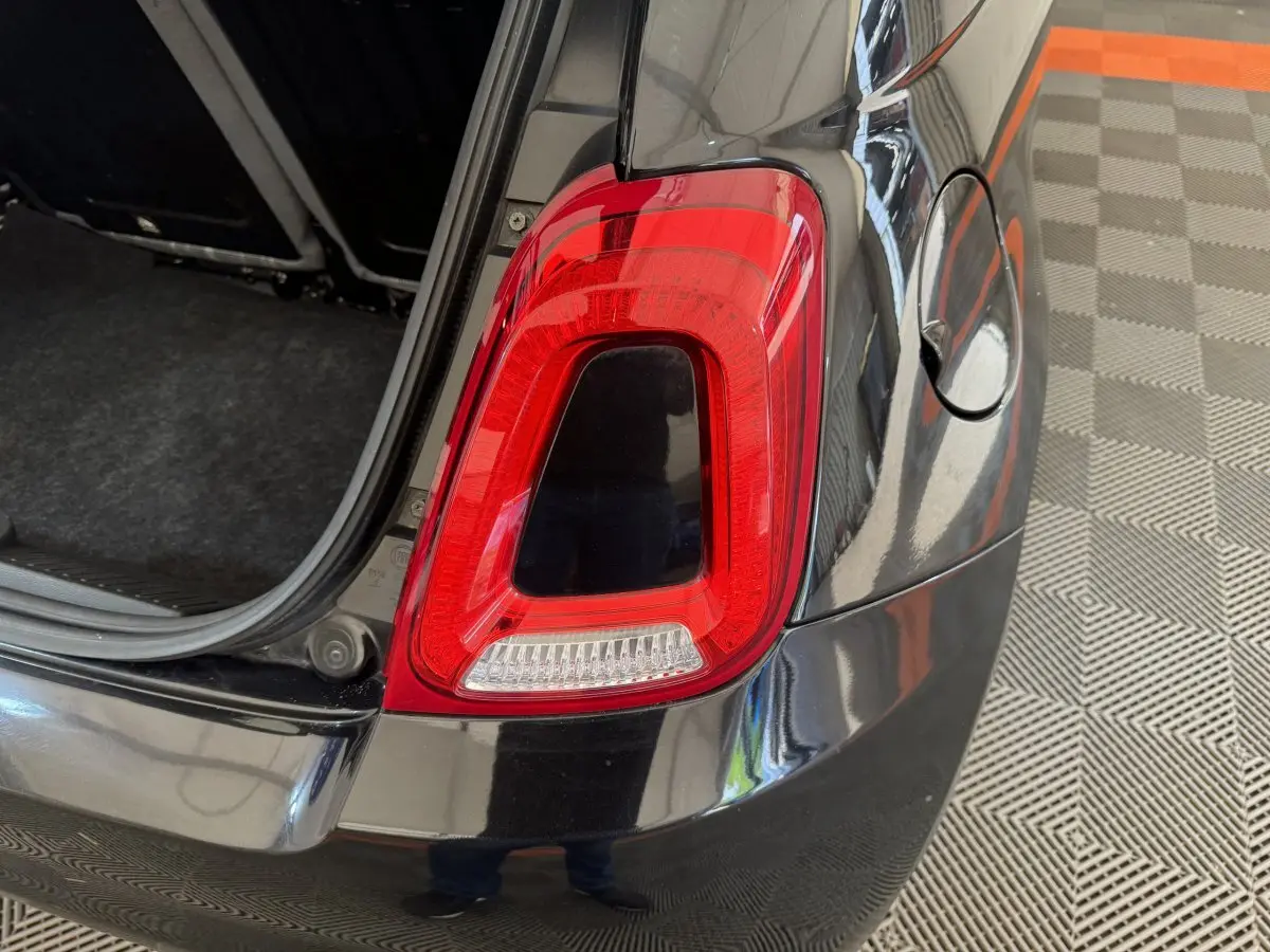 Gros plan sur le feu arrière droit rouge d'une Fiat 500 noire avec coffre ouvert, sur sol à damier gris et orange.