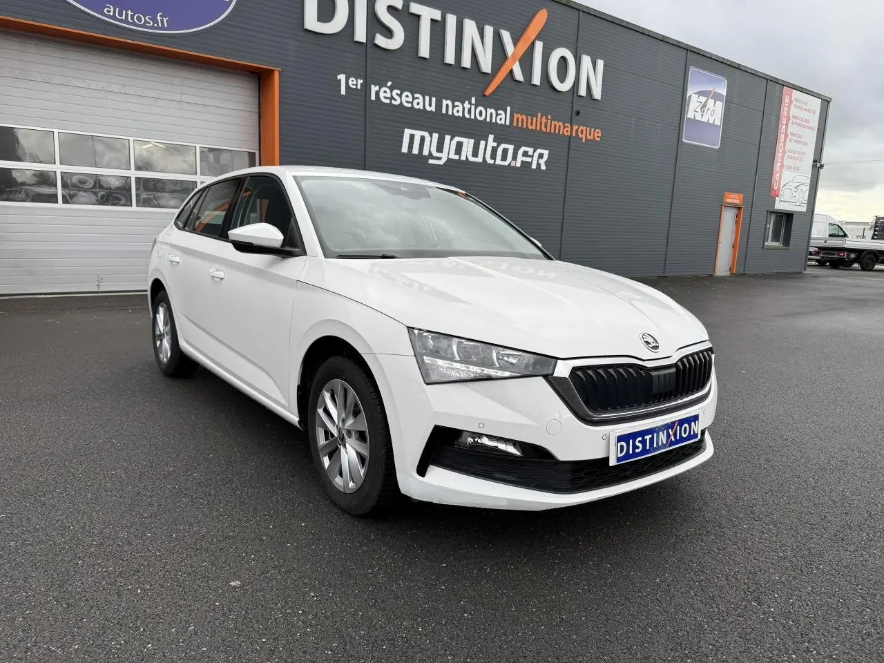 Vue 3/4 avant d'une Skoda Scala blanche 2023 avec jantes alliage argent devant un bâtiment gris.