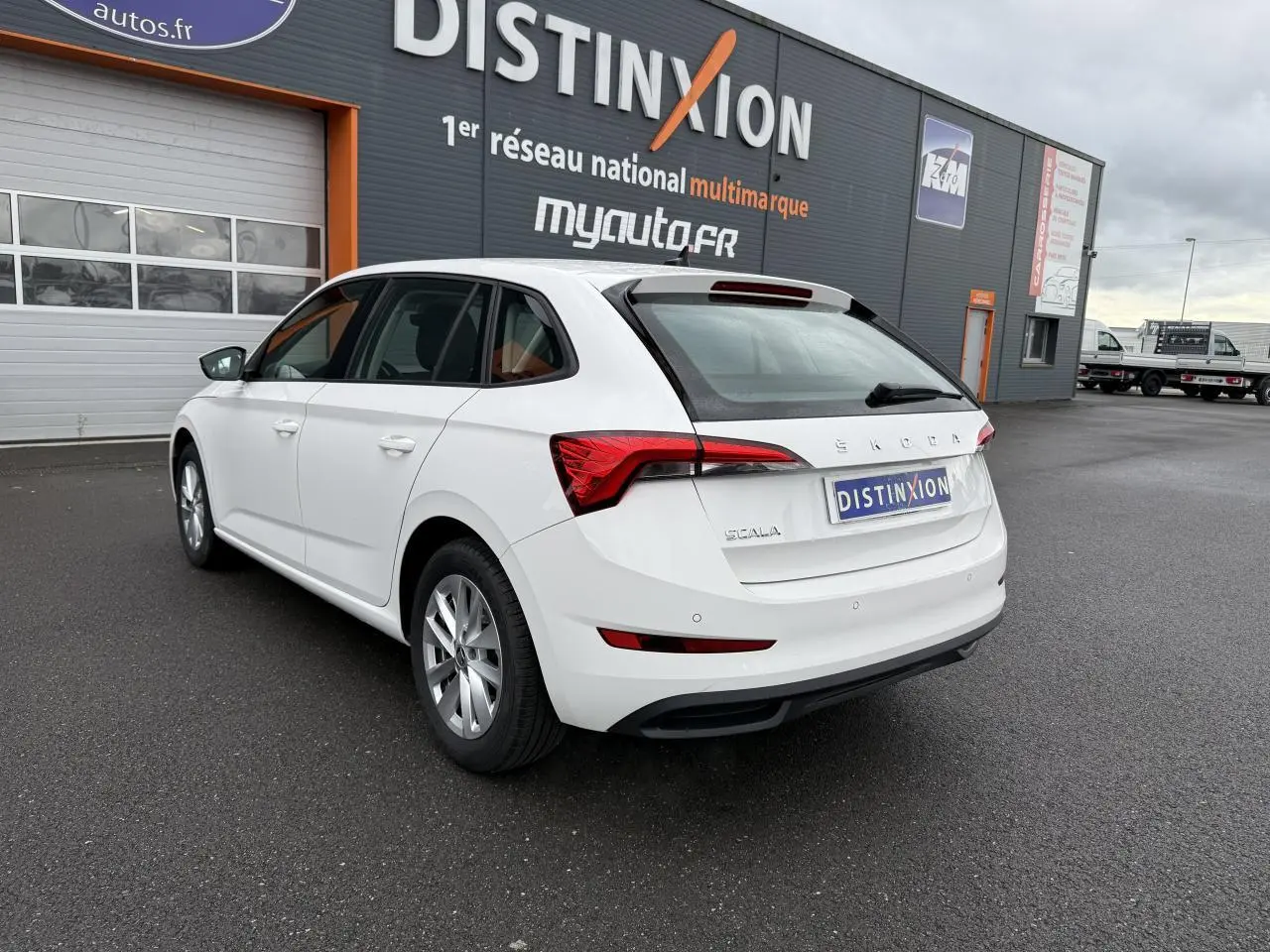 Vue 3/4 arrière droite d'une Skoda Scala blanche 2023 avec jantes alliage et feux arrière distinctifs, stationnée devant un bâtiment.