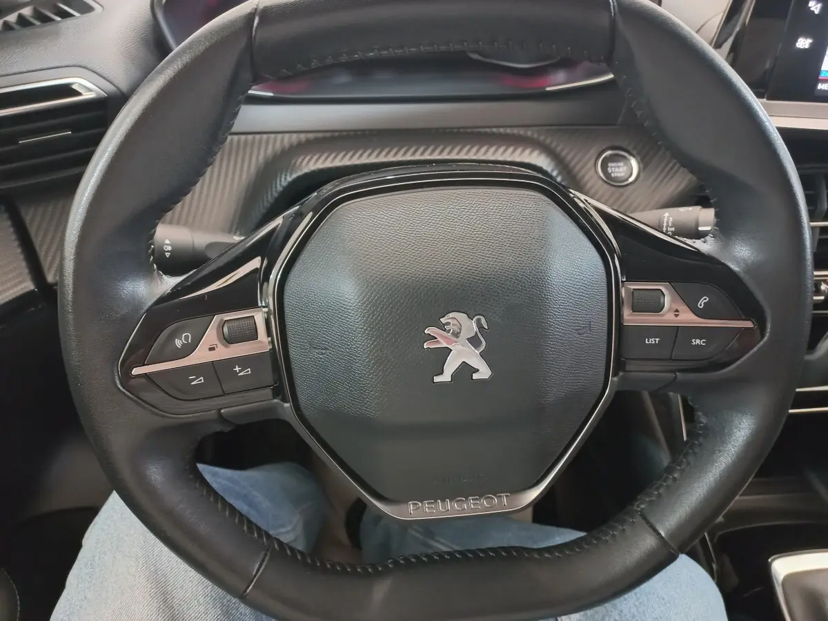 Vue rapprochée du volant cuir noir de la Peugeot 208 PureTech 100 S&S Allure avec commandes intégrées et logo lion central.