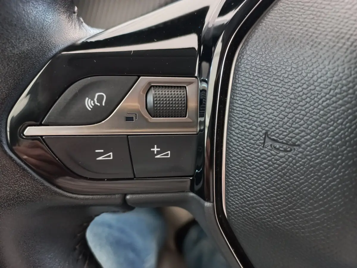 Gros plan sur les commandes de volume et de reconnaissance vocale sur le volant cuir noir d'une Peugeot 208 blanche.