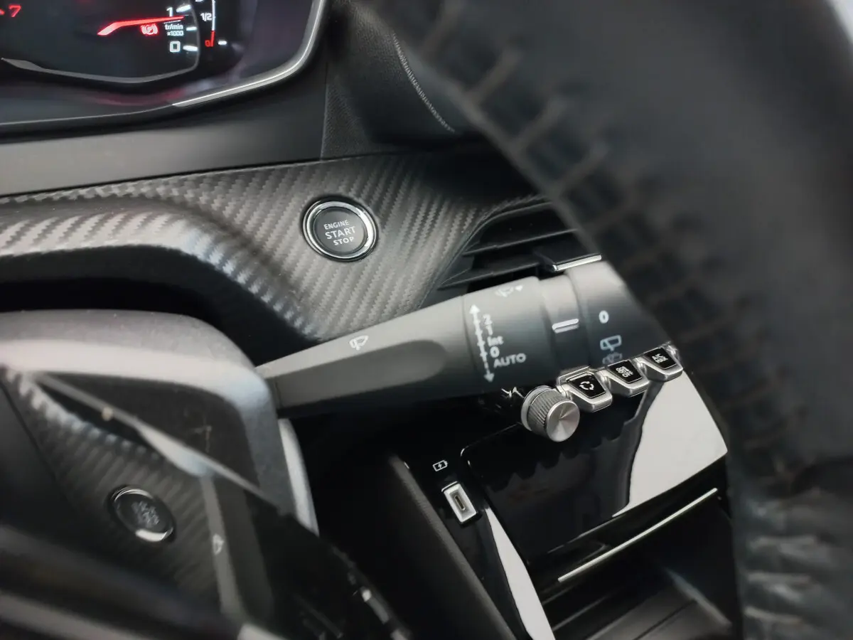 Gros plan sur le tableau de bord carbone et le bouton start/stop d'une Peugeot 208 blanche, version PureTech 100 S&S Allure.