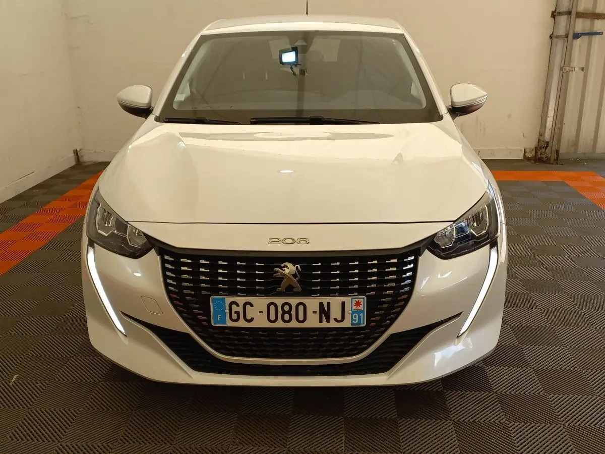 Vue frontale d'une Peugeot 208 blanche avec phares LED et calandre noire distinctive dans un garage.