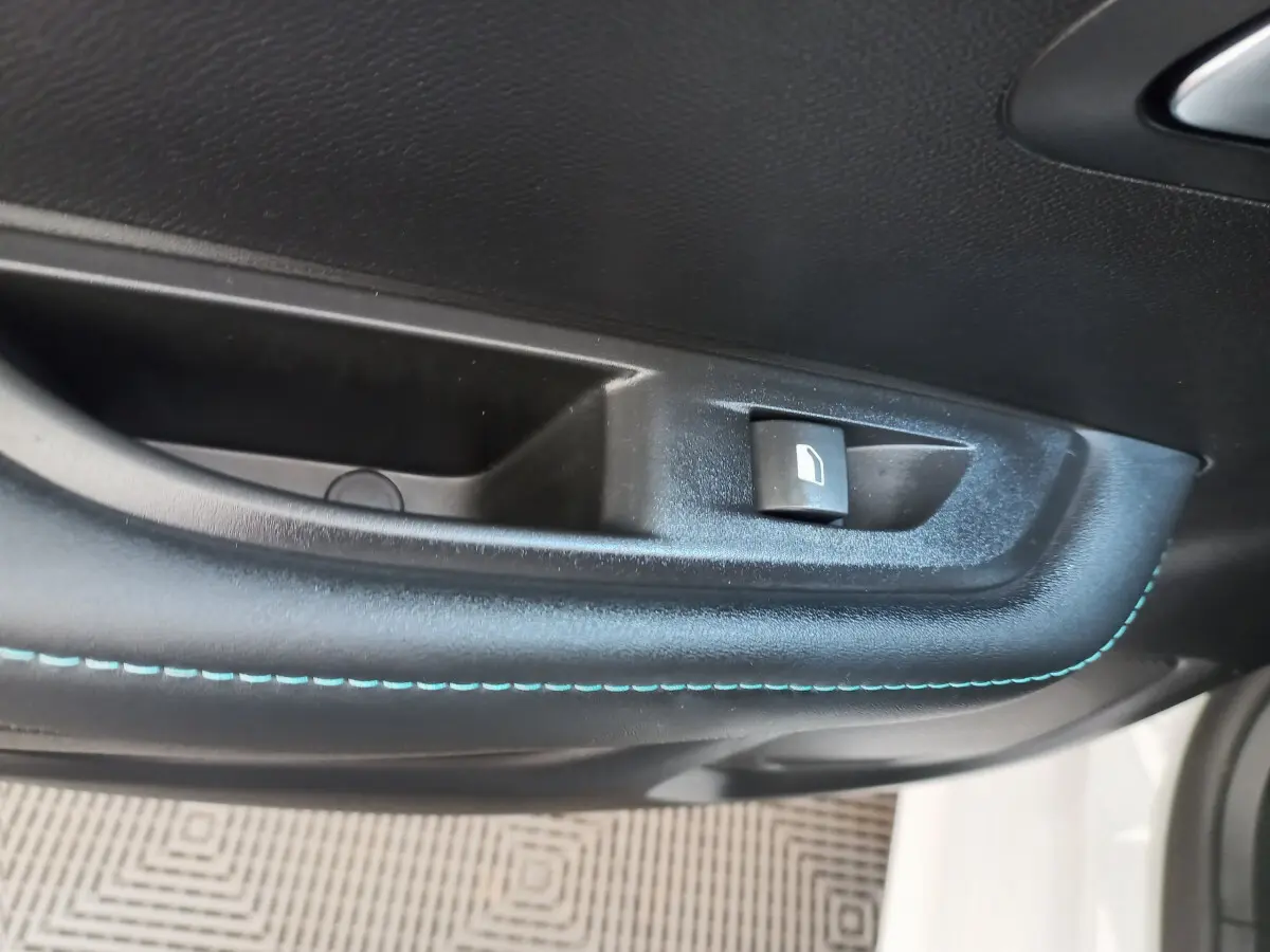 Gros plan sur la commande de lève-vitre côté gauche avec surpiqûres bleues dans l'habitacle d'une Peugeot 208 blanche.