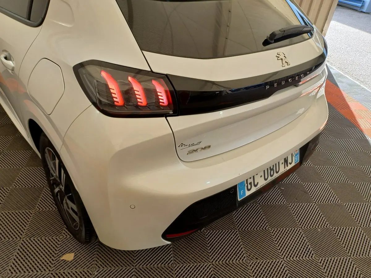 Vue 3/4 arrière droite d'une Peugeot 208 PureTech 100 S&S Allure blanche avec feux arrière LED distinctifs et logo Peugeot noir.