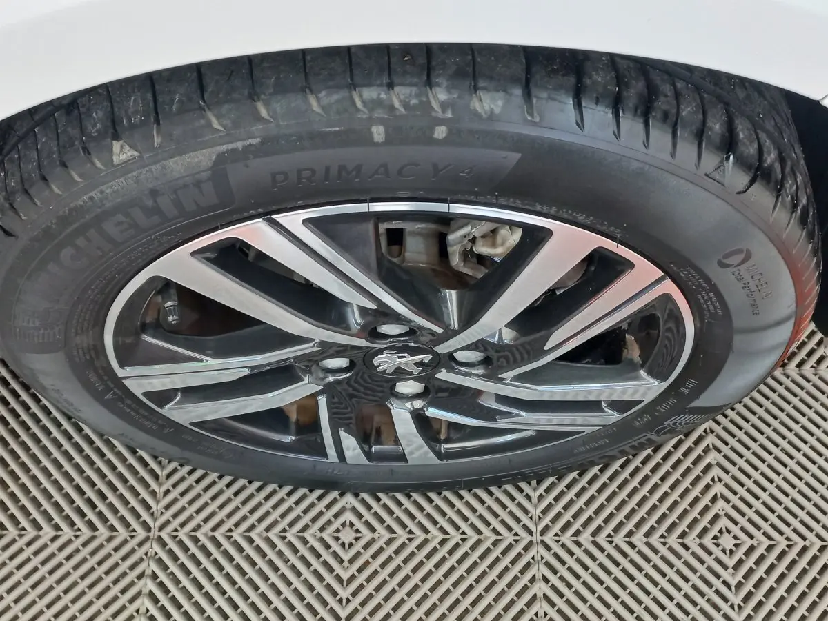 Gros plan sur la roue avant droite d'une Peugeot 208 blanche, mettant en valeur la jante alliage noire et argentée avec logo lion.