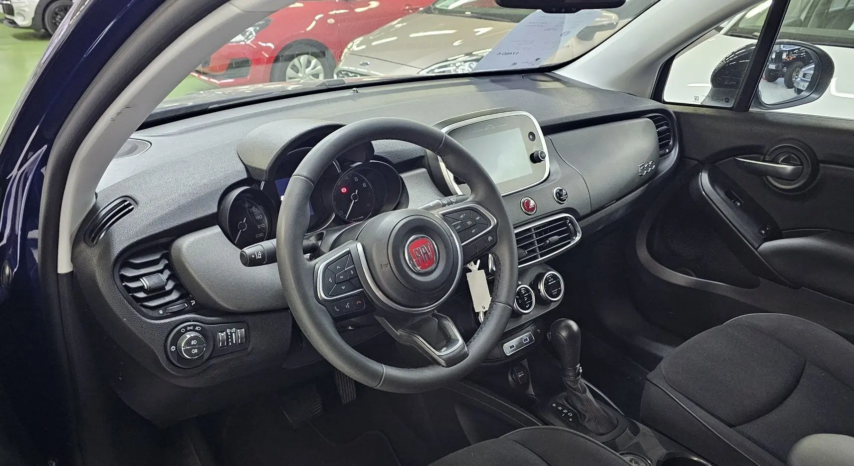Intérieur de la FIAT 500X 2024 en bleu, vue côté conducteur montrant le volant, tableau de bord et console centrale.