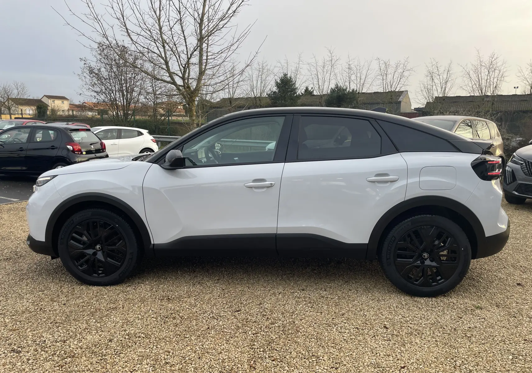 Profil côté gauche d'une Citroën C4 2025 blanc Okenite avec toit noir Perla et jantes noires sur parking extérieur.
