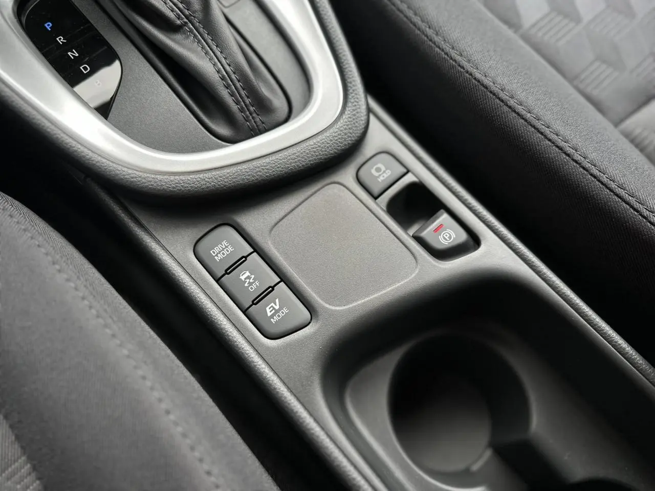 Gros plan sur la console centrale de la Toyota Yaris hybride 2025 avec boutons Drive Mode, frein électrique et sellerie tissu noir.