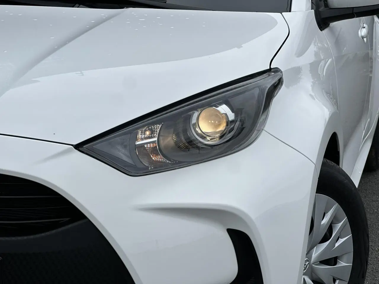 Gros plan sur le phare avant droit allumé d'une Toyota Yaris hybride blanche 2025, avec jante et aile visibles.
