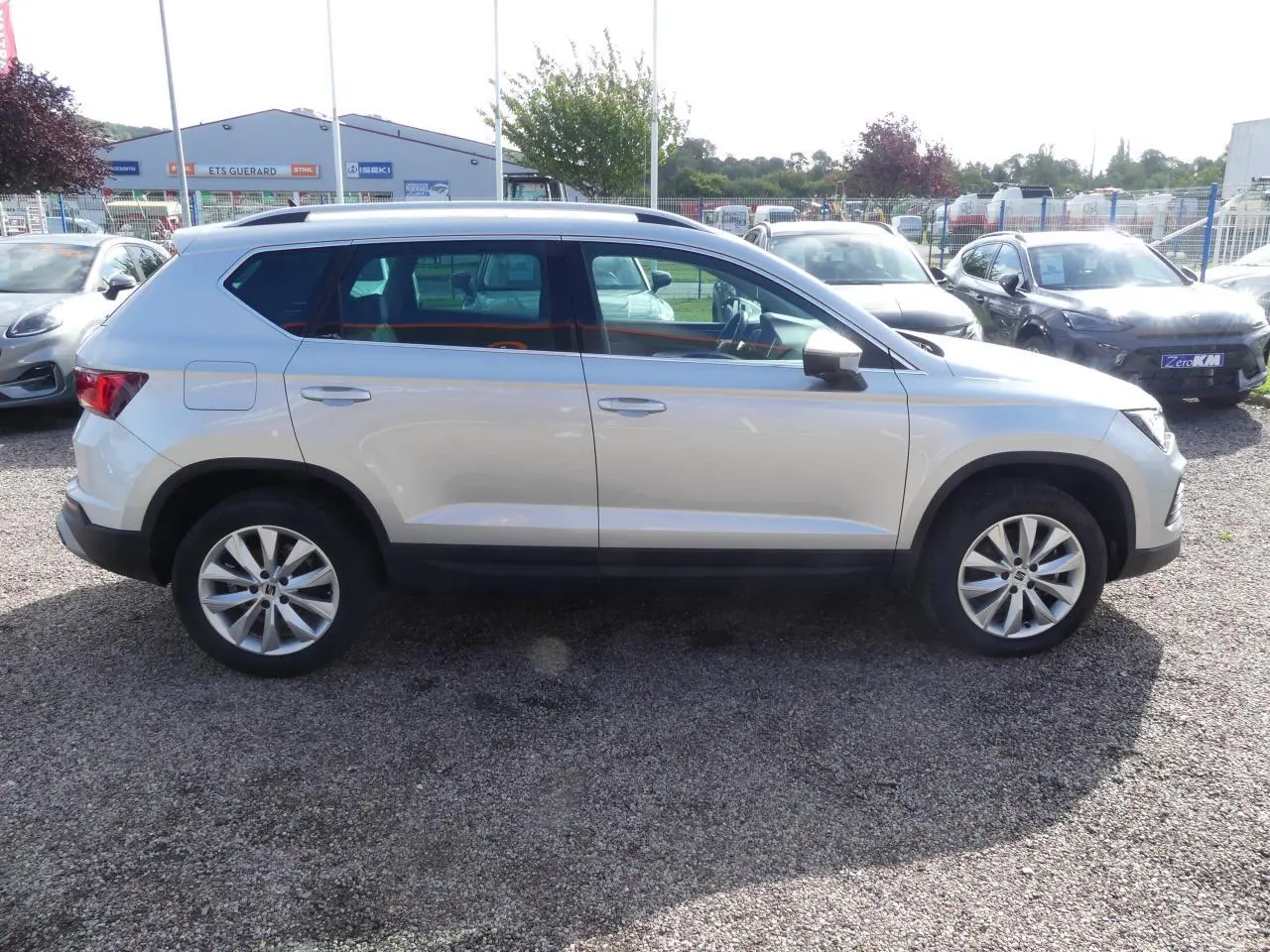 Vue de profil côté gauche d'un SEAT Ateca gris clair 2023 avec jantes alliage 17 pouces et barres de toit chromées.