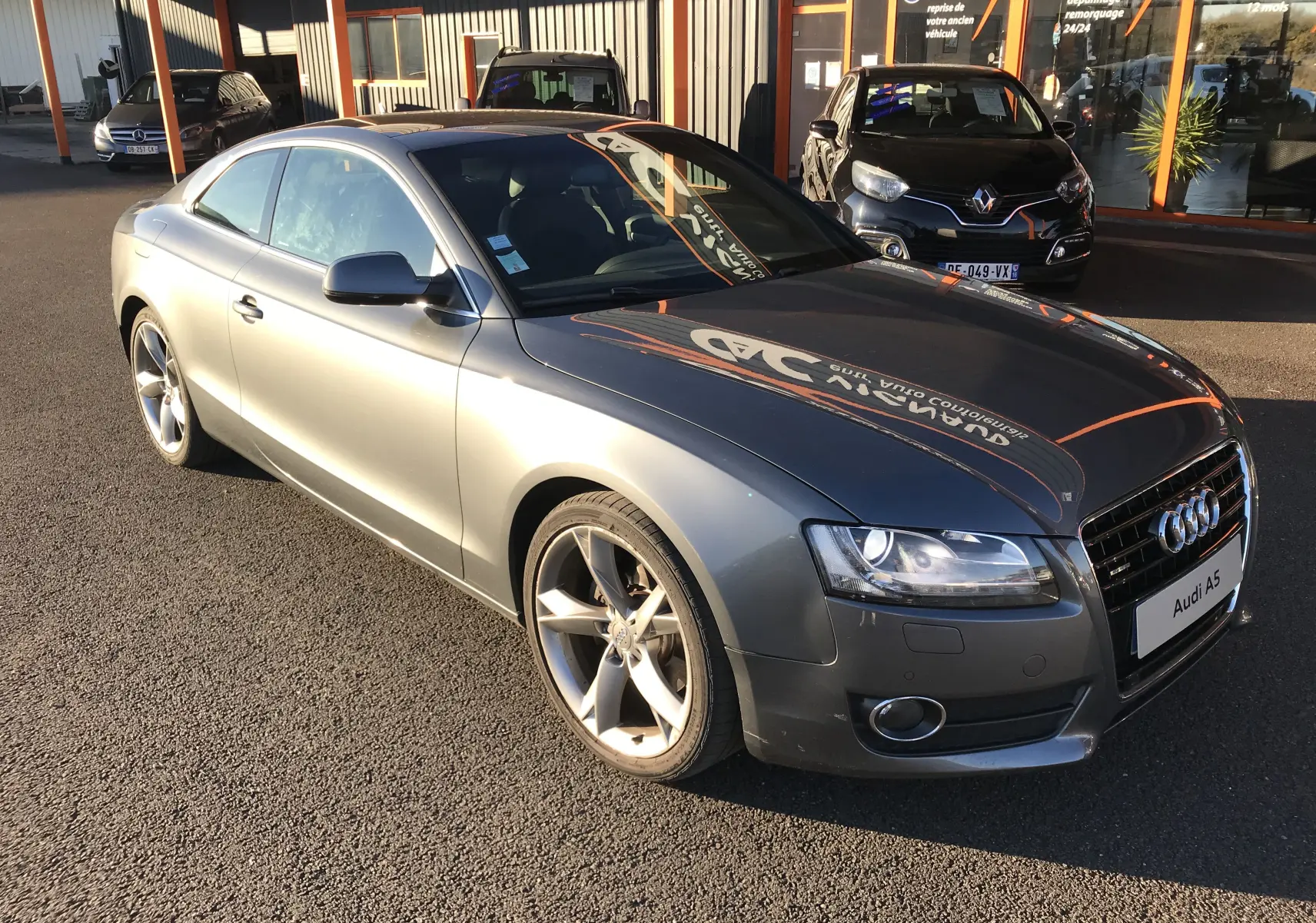 Audi A5 Coupé gris dauphin vu de 3/4 avant droit, avec jantes alliage et phares allumés.