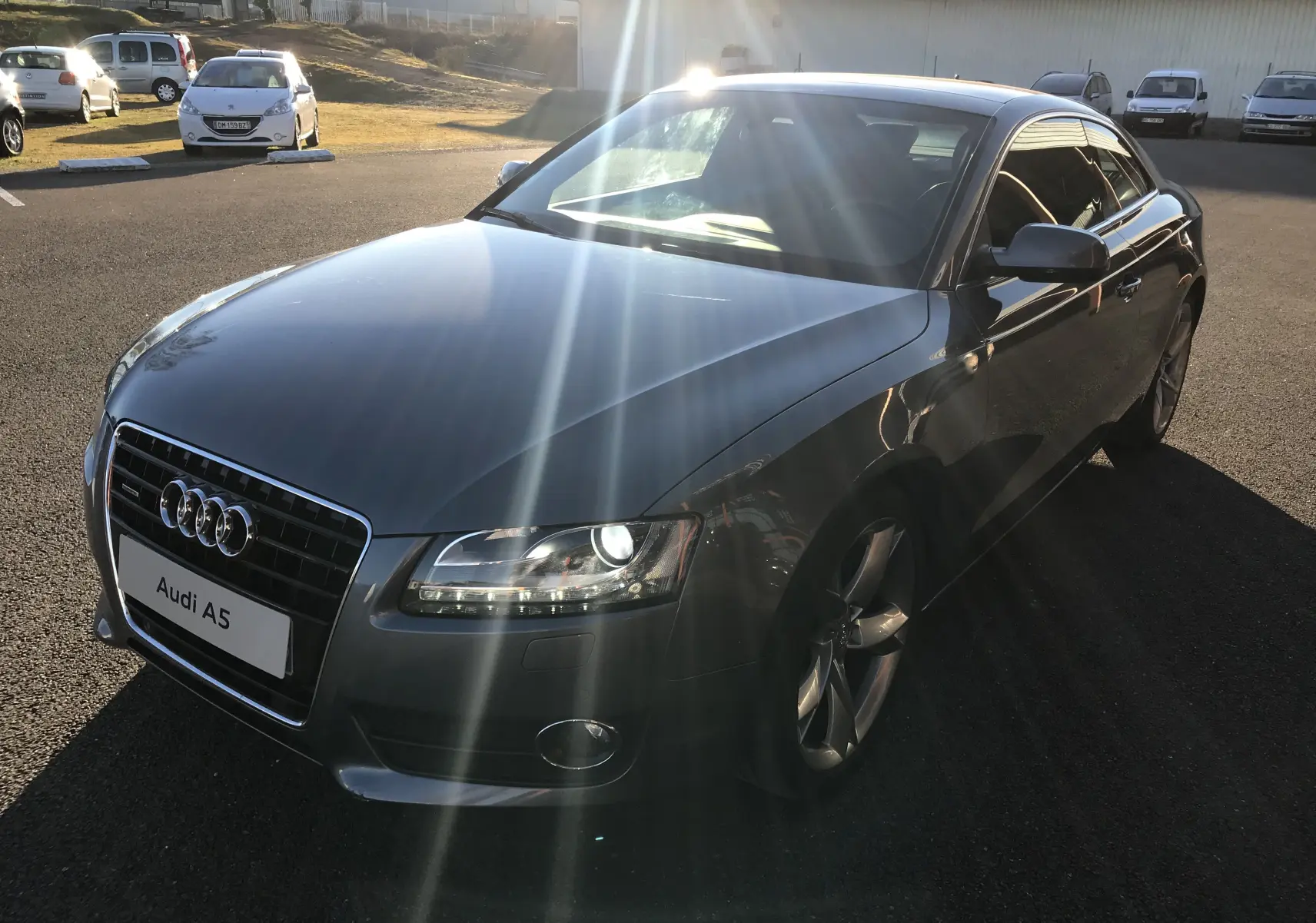 Audi A5 Coupé gris dauphin vu en 3/4 avant droit avec phares allumés et jantes alliage cinq branches.