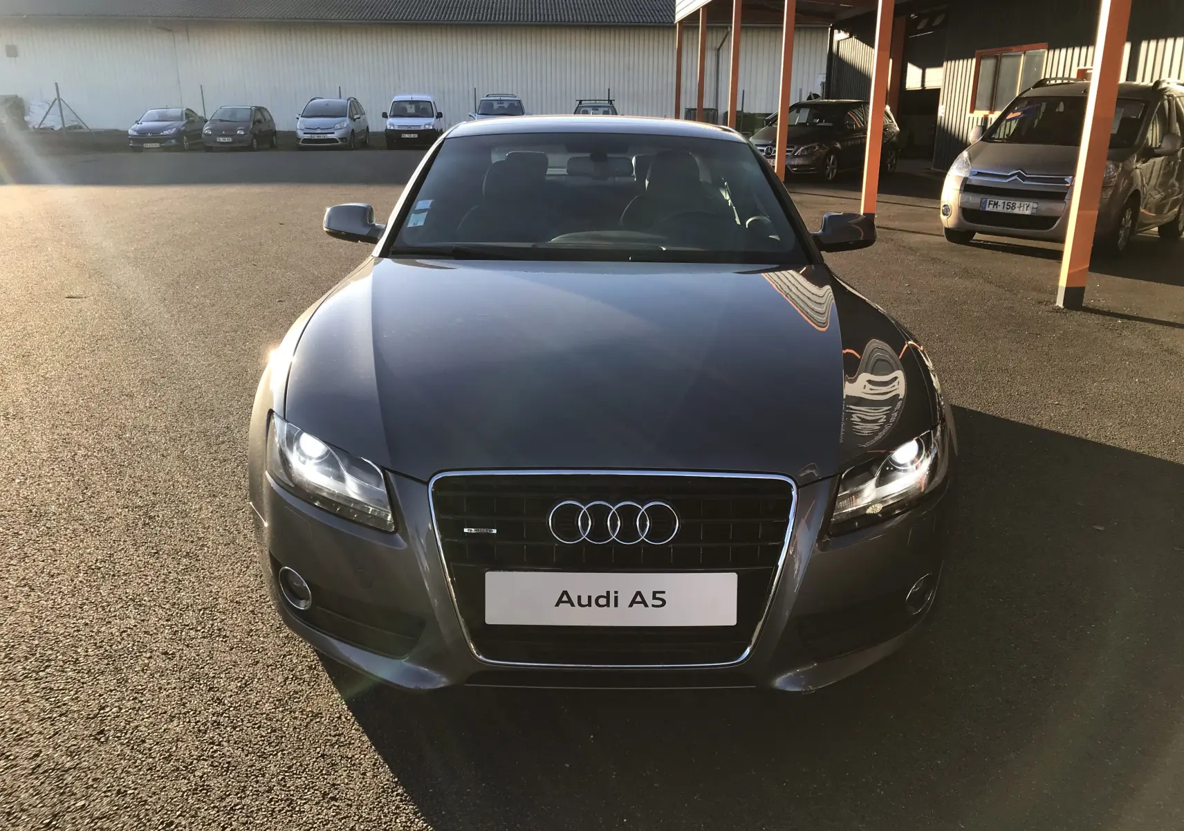 Vue frontale d'une Audi A5 gris dauphin avec phares allumés, garée en extérieur sous un soleil bas.