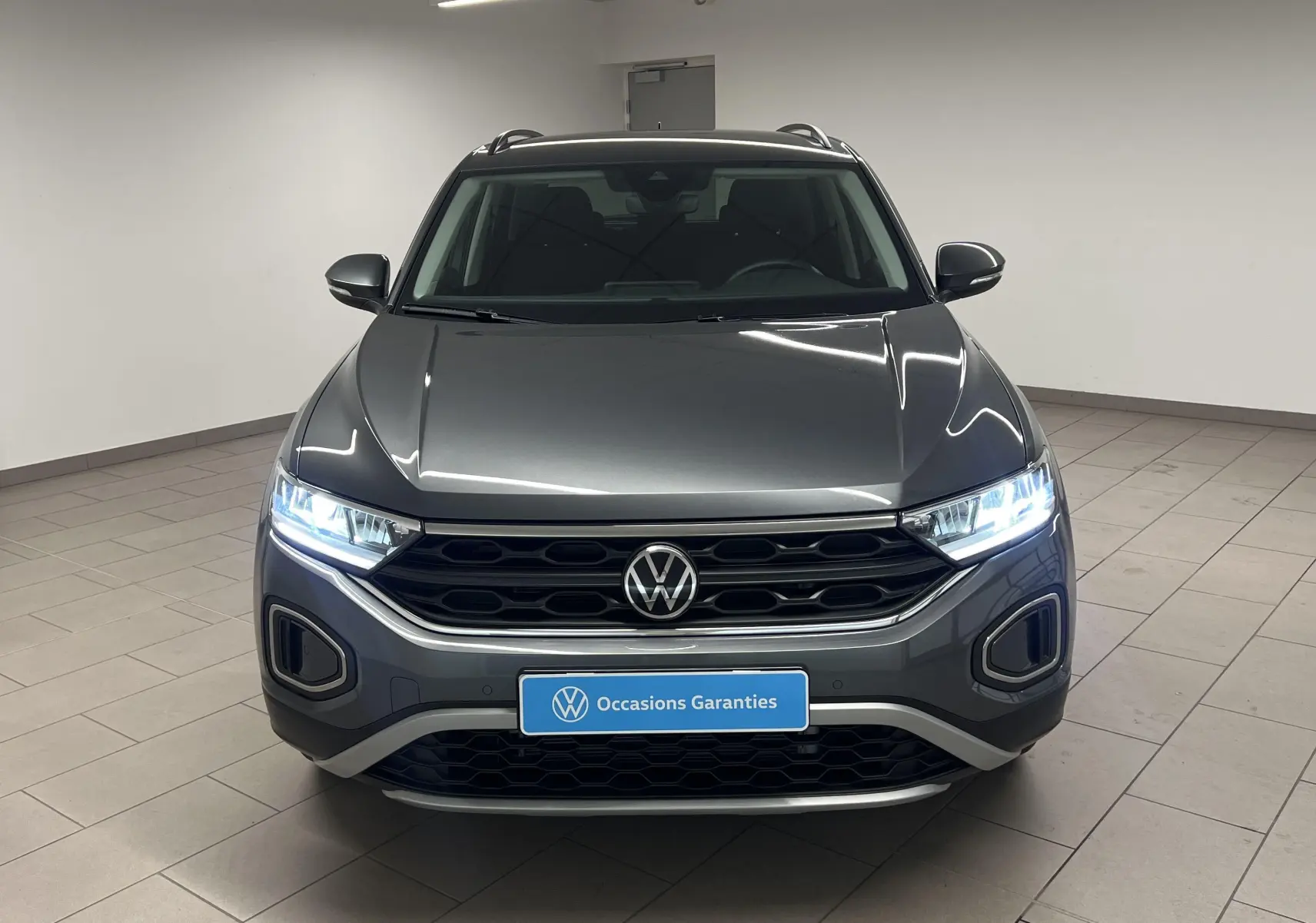 Volkswagen T-Roc gris Indium vu de face avec phares allumés et calandre noire distinctive.