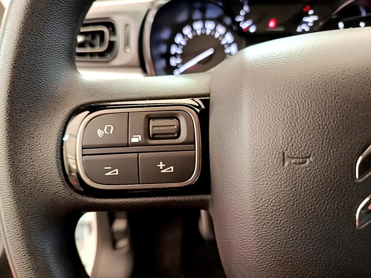 Gros plan sur les commandes gauche du volant noir d'une Citroën C3 blanche, avec tableau de bord flou en arrière-plan.