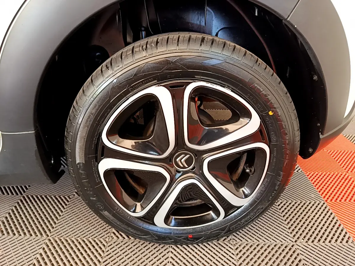 Gros plan sur la roue arrière droite d'une Citroën C3 blanche, avec passage de roue noir mat et sol à damier rouge et gris.