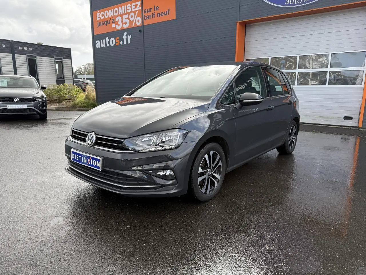 Volkswagen Golf Sportsvan gris Urano vue 3/4 avant droit sur parking devant un bâtiment commercial.