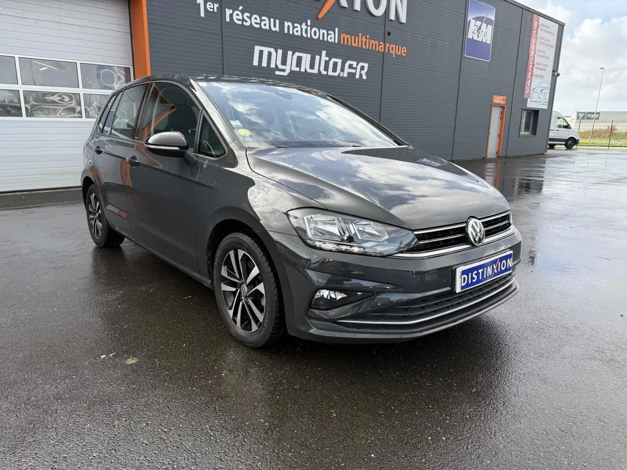 Volkswagen Golf Sportsvan gris Urano vue 3/4 avant droit sur sol humide devant un bâtiment commercial.