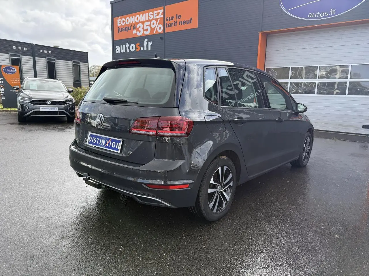Vue 3/4 arrière droite d'une Volkswagen Golf Sportsvan gris Urano stationnée devant un garage autos.fr.