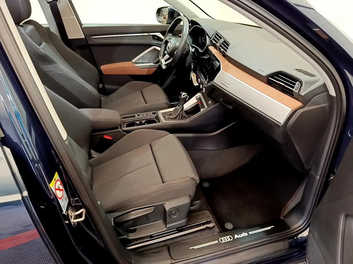 Intérieur avant droit de l'Audi Q3 bleu 2020, sièges gris, tableau de bord avec insert bois et volant cuir multifonctions.