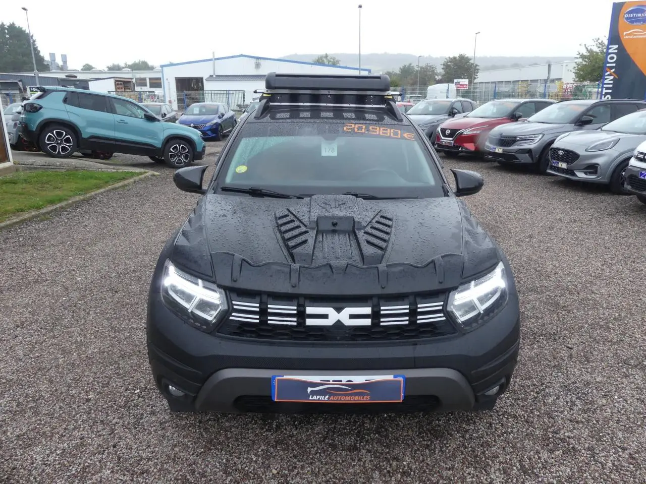 Vue avant d'un Dacia Duster noir 2023 avec covering intégral et galerie de toit sur un parking humide.