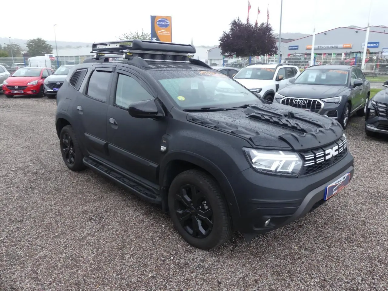 Dacia Duster noir 2023 en 3/4 avant droit avec galerie de toit, marchepieds et jantes noires sur parking.