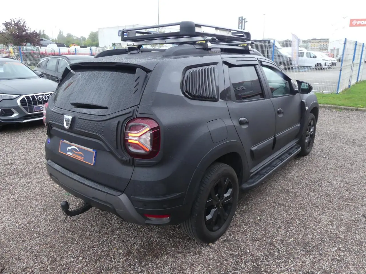 Vue 3/4 arrière droite du Dacia Duster noir 2023 avec galerie de toit, attelage fixe et marchepieds.